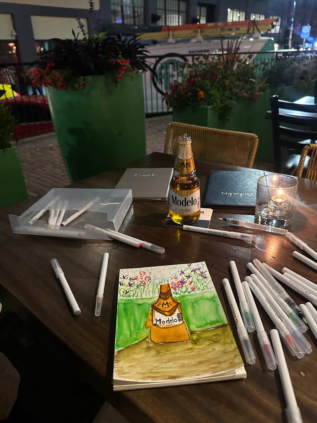 Table with a bottle of Modelo beer, a drawing of the same bottle, and art supplies in an outdoor setting.