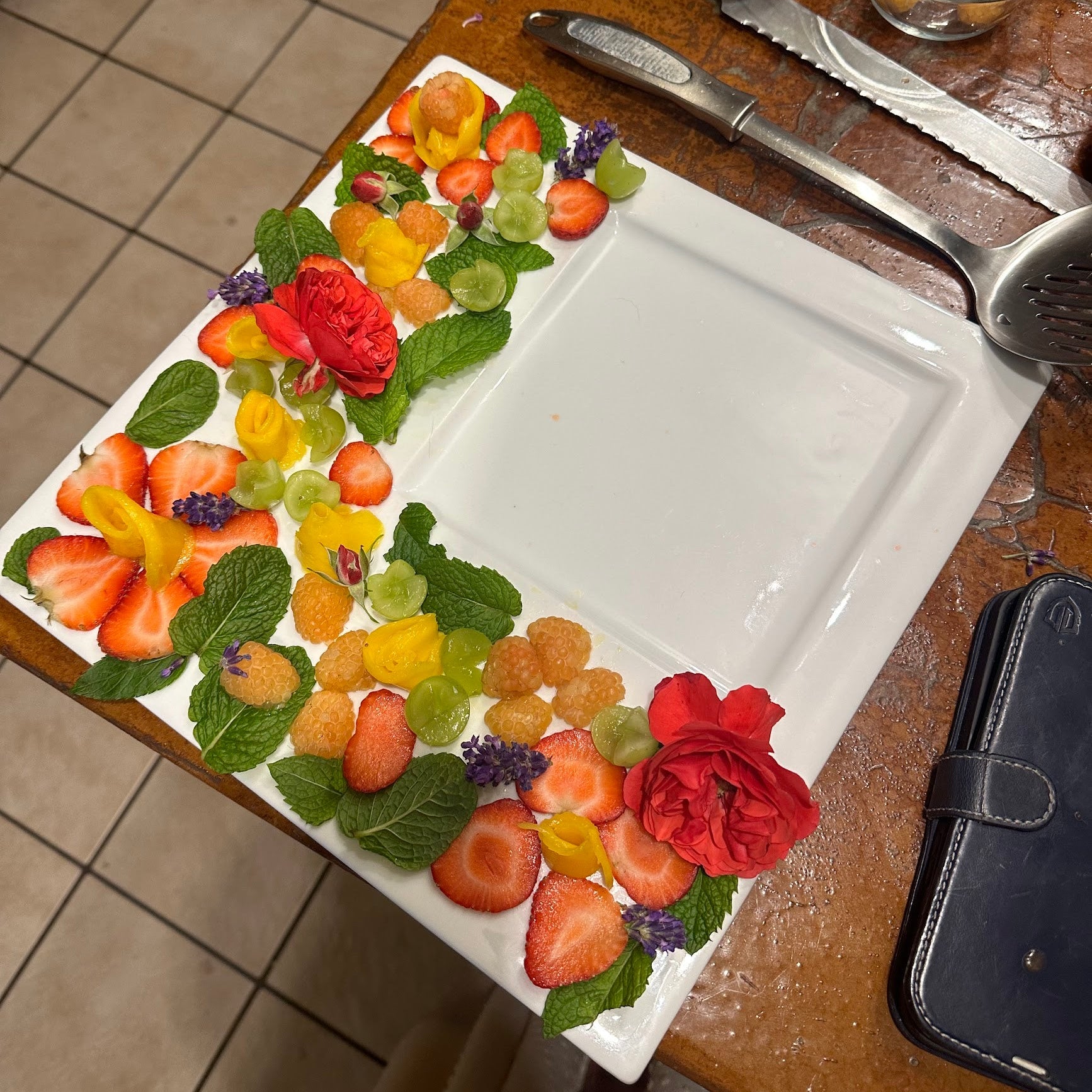 Decorative fruit platter on a white tray with kitchen utensils and a phone in the background.