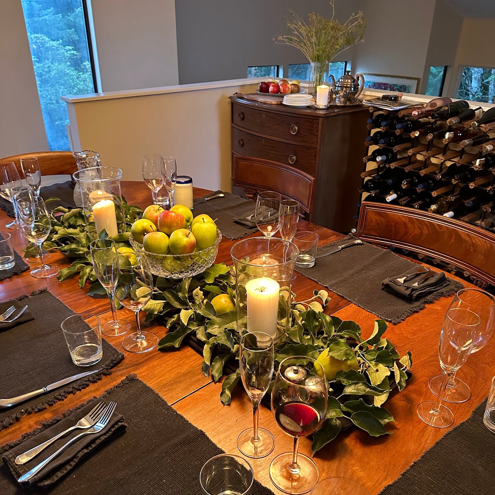 Dining table set for a meal with wine glasses, candles, and fruit, in a room with wine bottles and a window.