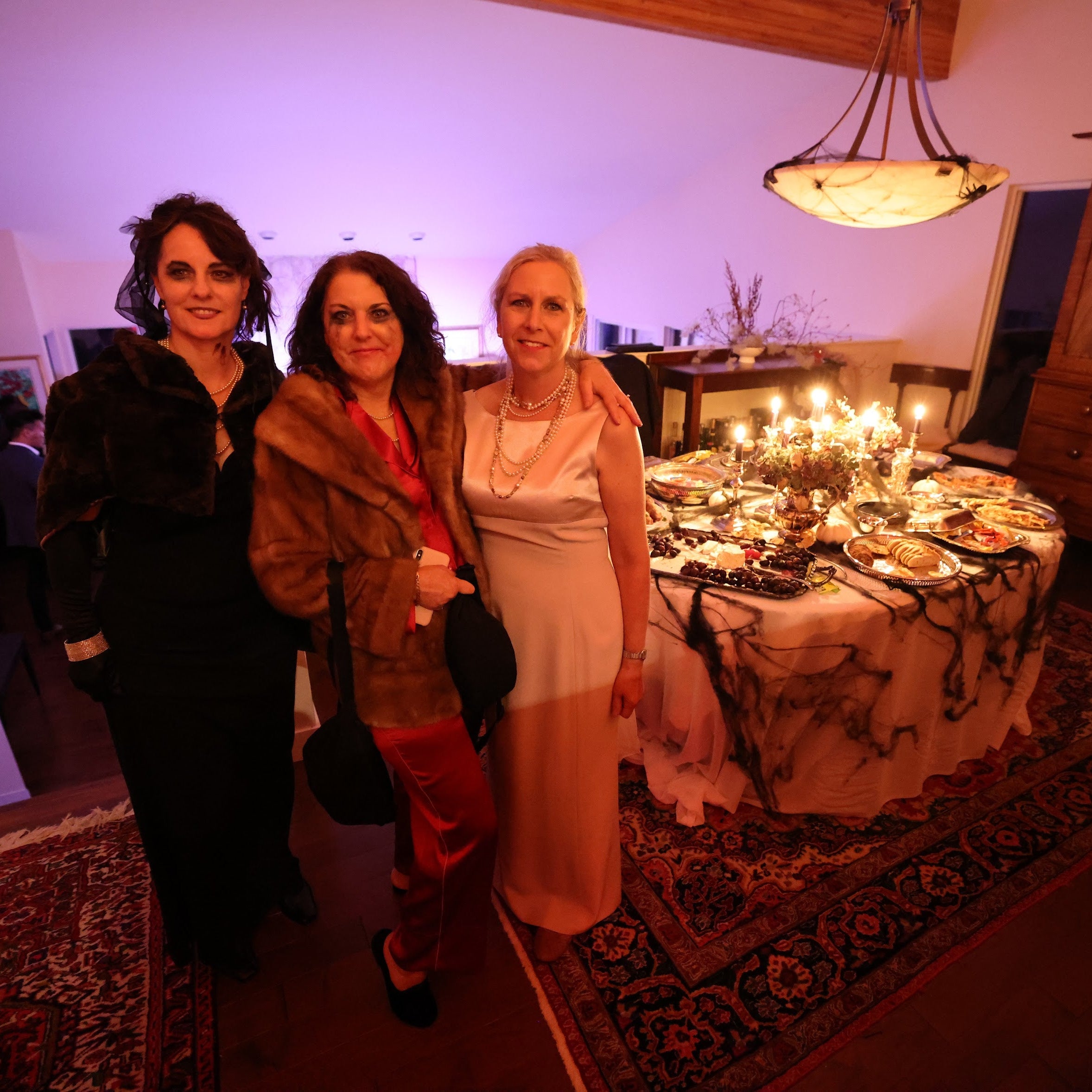 Three women posing in a room with a decorated table and chandelier.