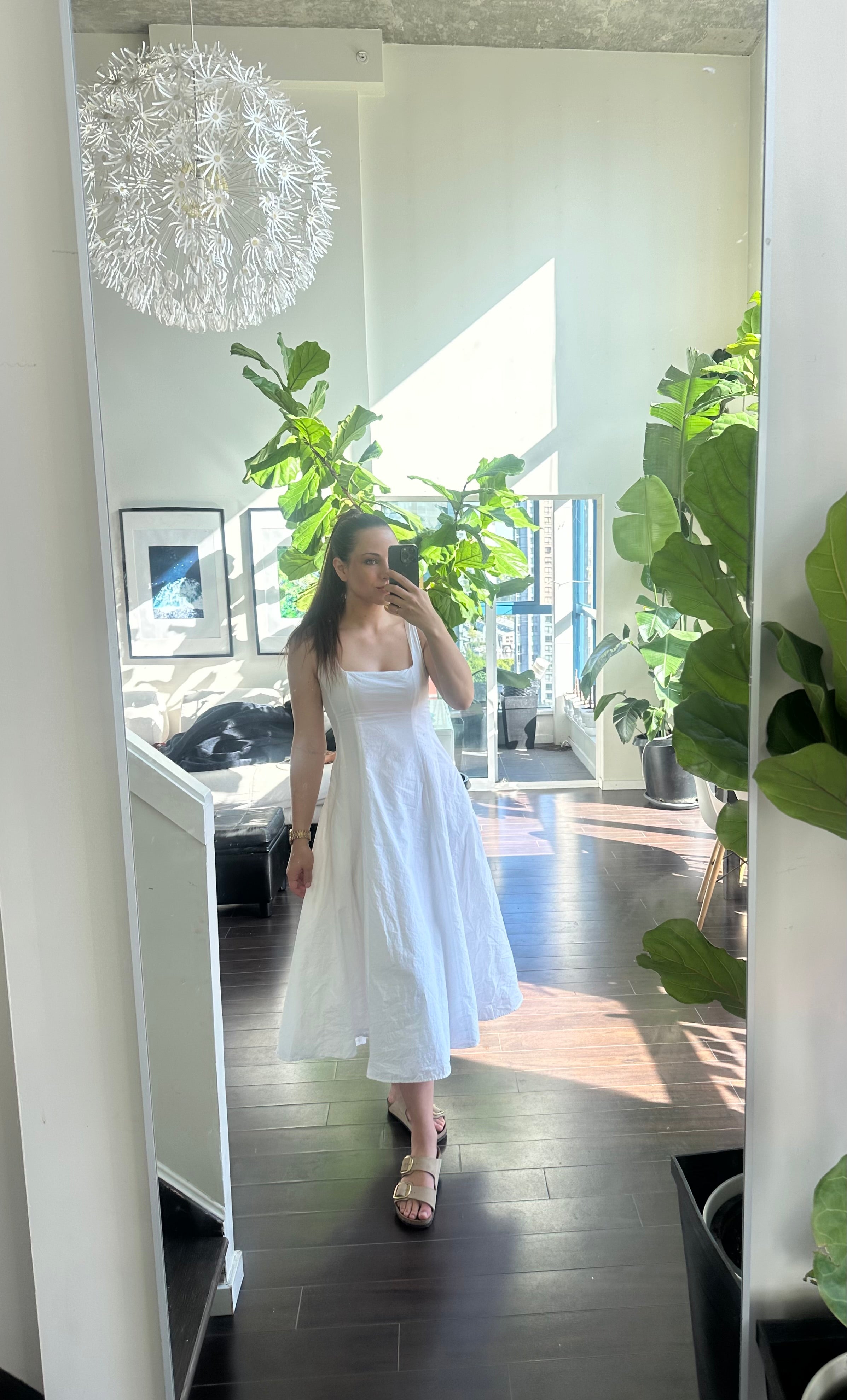 Woman in a white dress taking a mirror selfie in a bright room with plants and furniture.