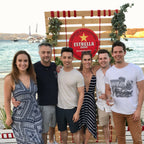 Group of six people posing together on a wooden deck by a waterfront, with a prominent Estrella beer sign in the background.