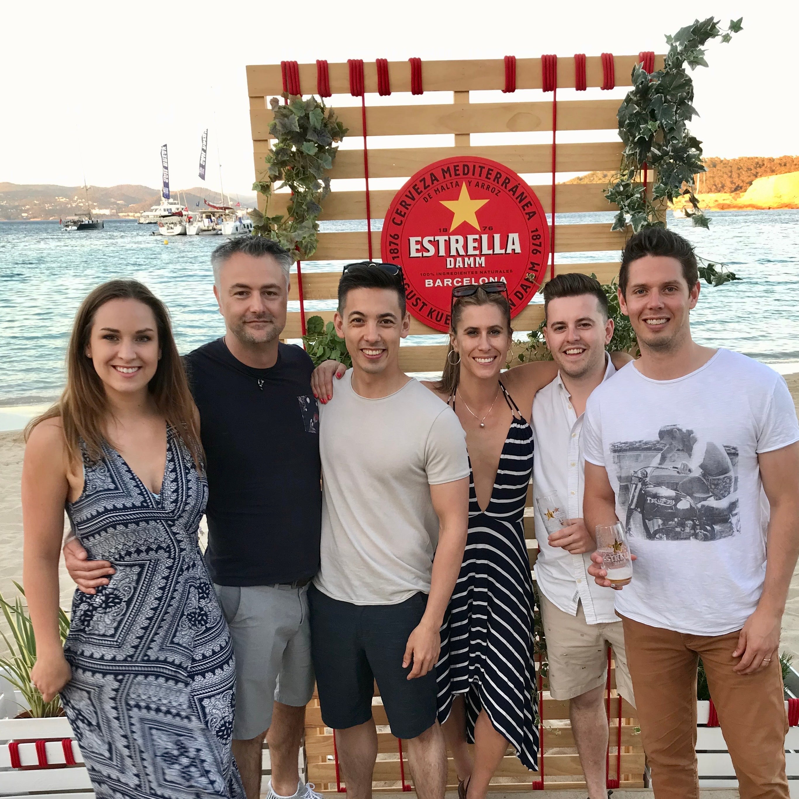 Group of six people posing together on a wooden deck by a waterfront, with a prominent Estrella beer sign in the background.