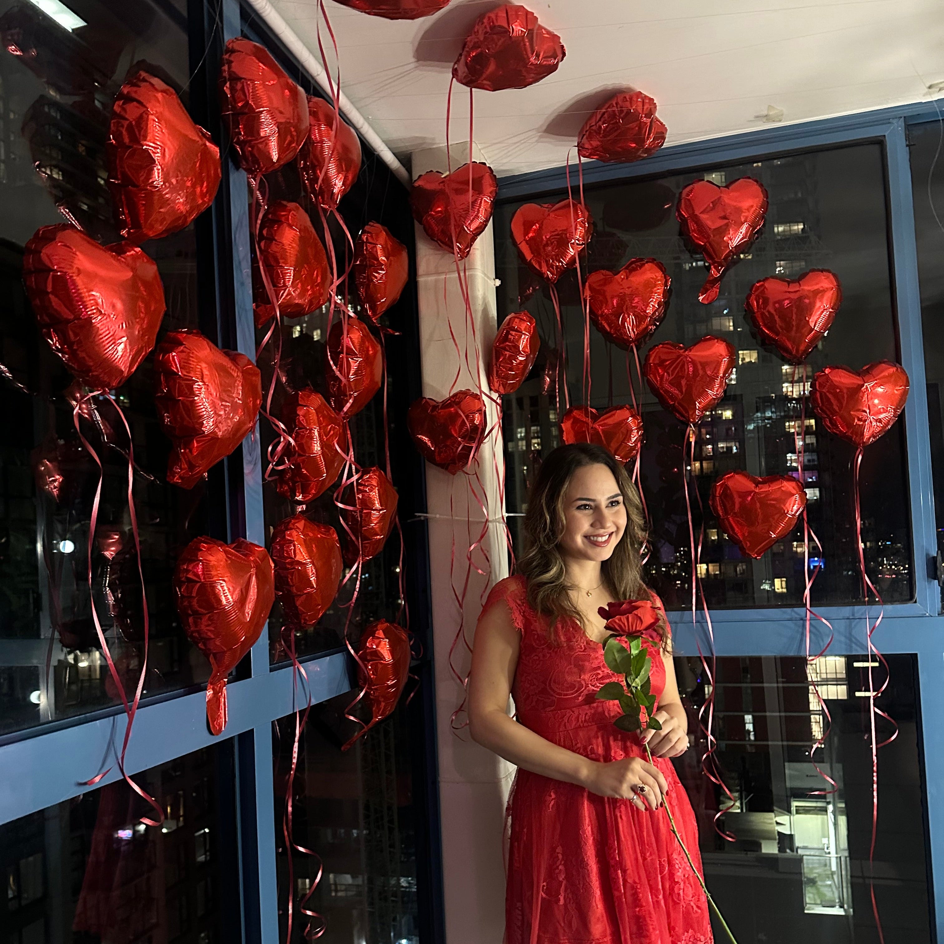 Woman in a red dress surrounded by heart-shaped balloons inside a glass display case.