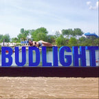 Large blue sign with 'LAVIEBUDLIGHT' on a beach with people and boats in the background.