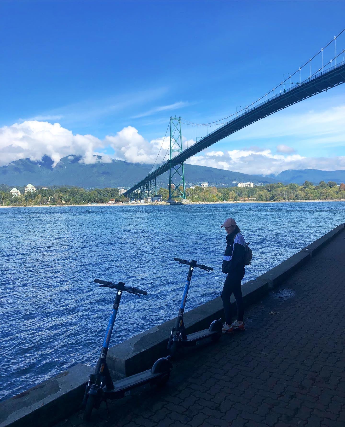 Person with electric scooters by a waterfront with a bridge and mountains in the background