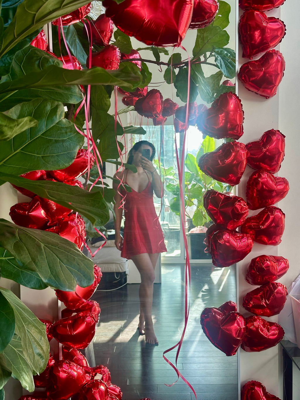 Person in a red dress standing in front of a decorative arrangement of red heart-shaped balloons and green leaves.