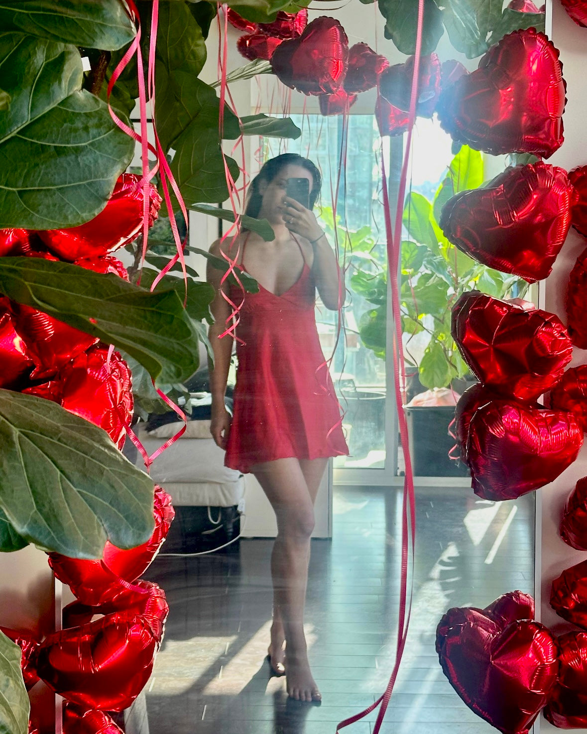Person in a red dress standing among red heart-shaped balloons and greenery.