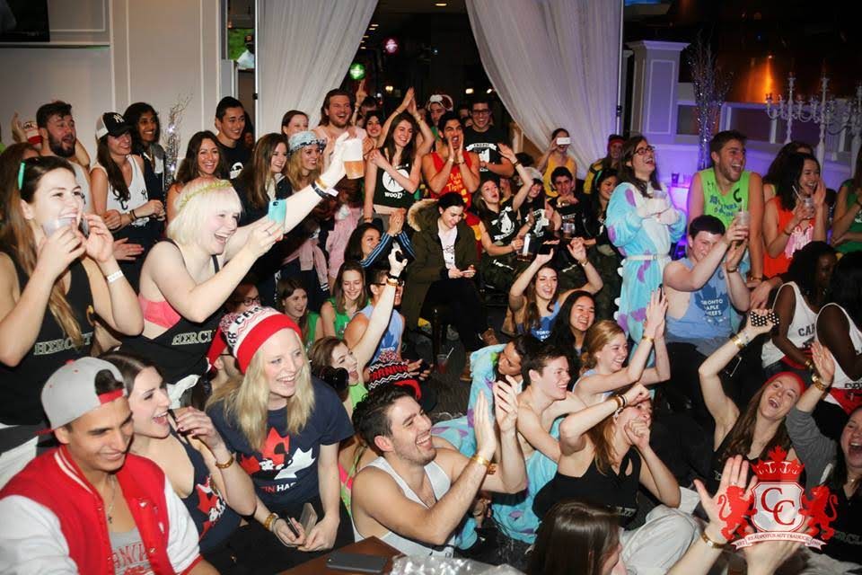 People at a party with raised hands, some holding drinks, in a dimly lit indoor setting.