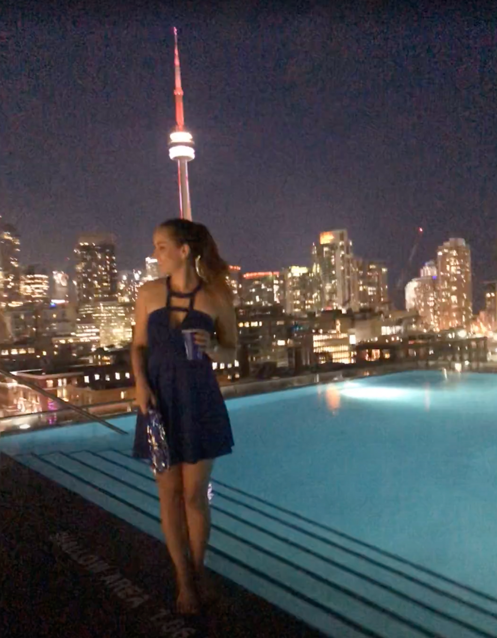 Woman in a blue dress standing by an infinity pool with a city skyline and CN Tower in the background.