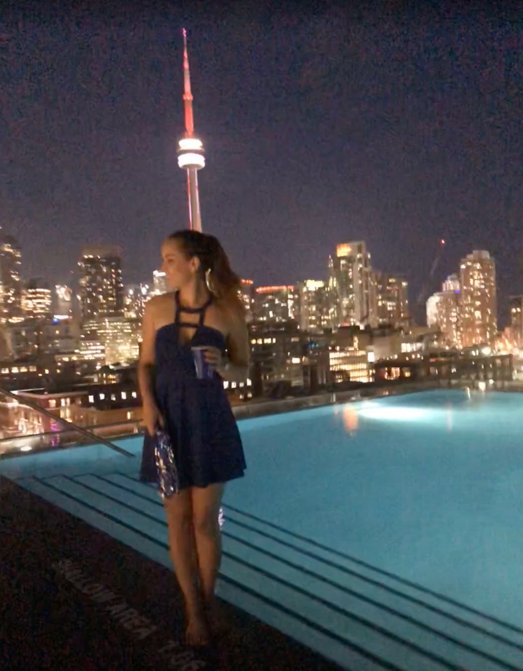Woman in a blue dress standing by an infinity pool with a city skyline and CN Tower in the background.