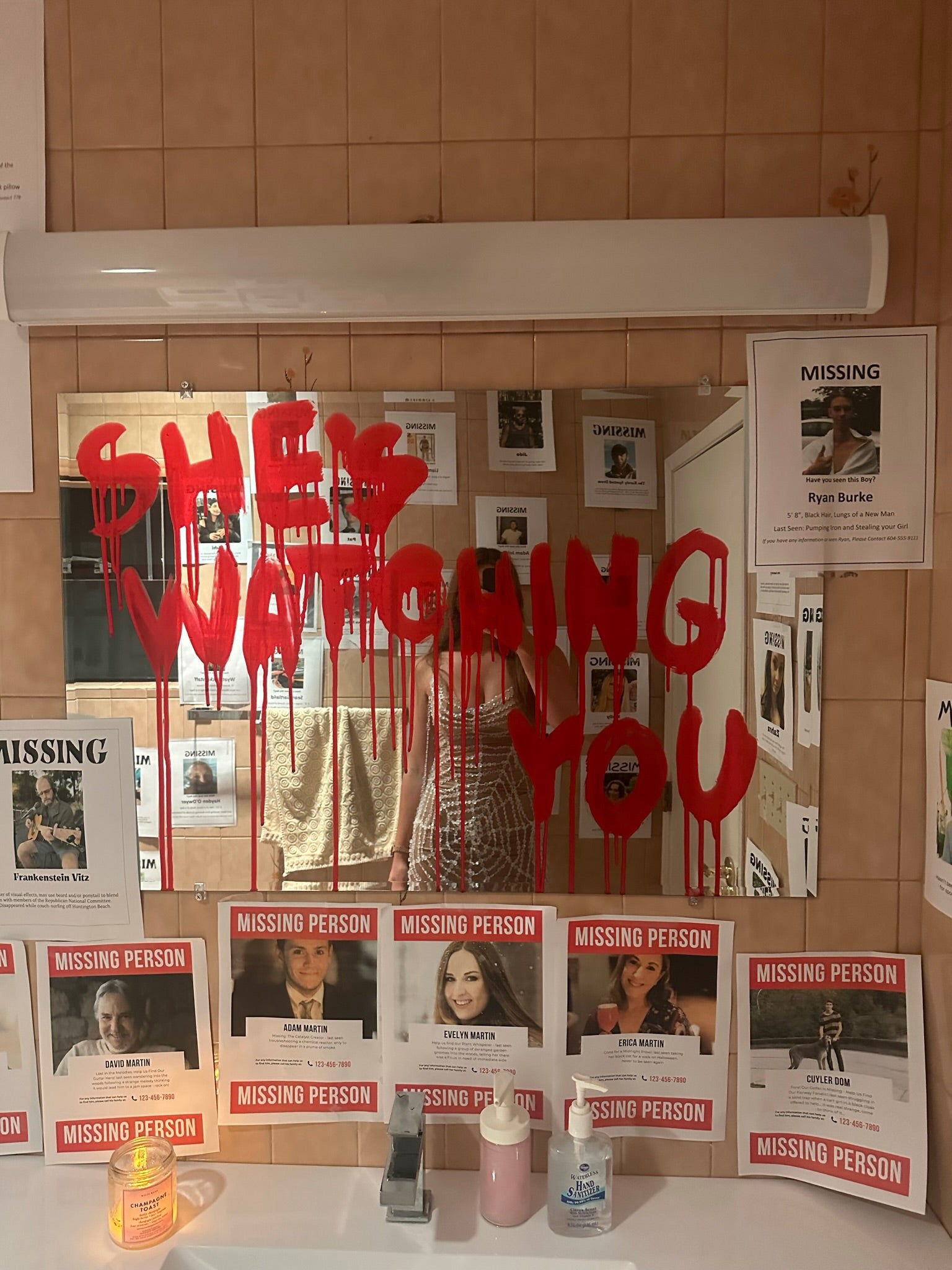 Party bathroom mirror reflection with missing person posters and red bloody paint on a mirror saying she’s watching you from the Blair witch party 
