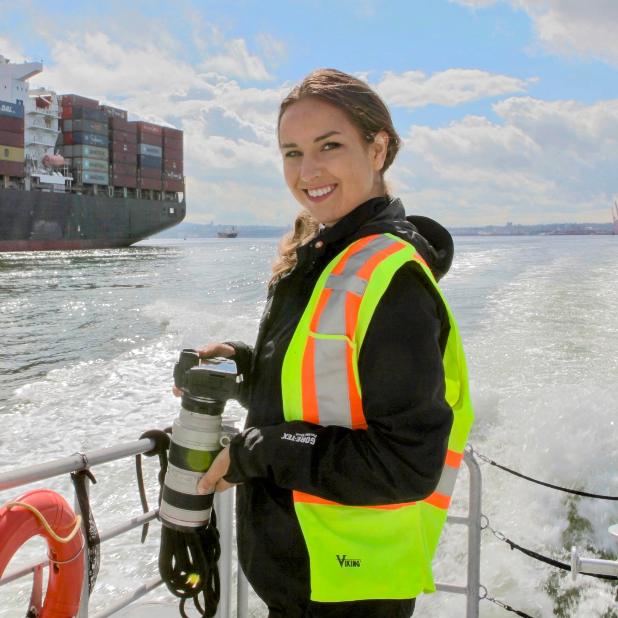 Person in a high-visibility vest standing on a boat with a large container ship in the background. Evelyn boating bc hero 