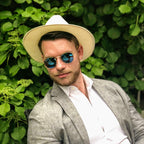 Man wearing a wide-brimmed hat and sunglasses, standing against a green leafy background