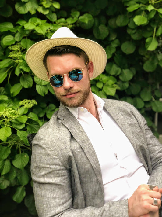 Man wearing a wide-brimmed hat and sunglasses, standing against a green leafy background