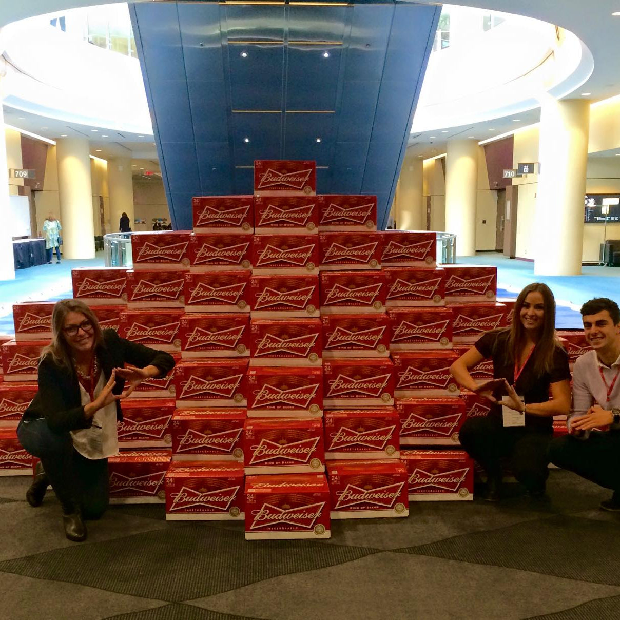 Three people posing with a large stack of red and white boxes in a spacious indoor area.