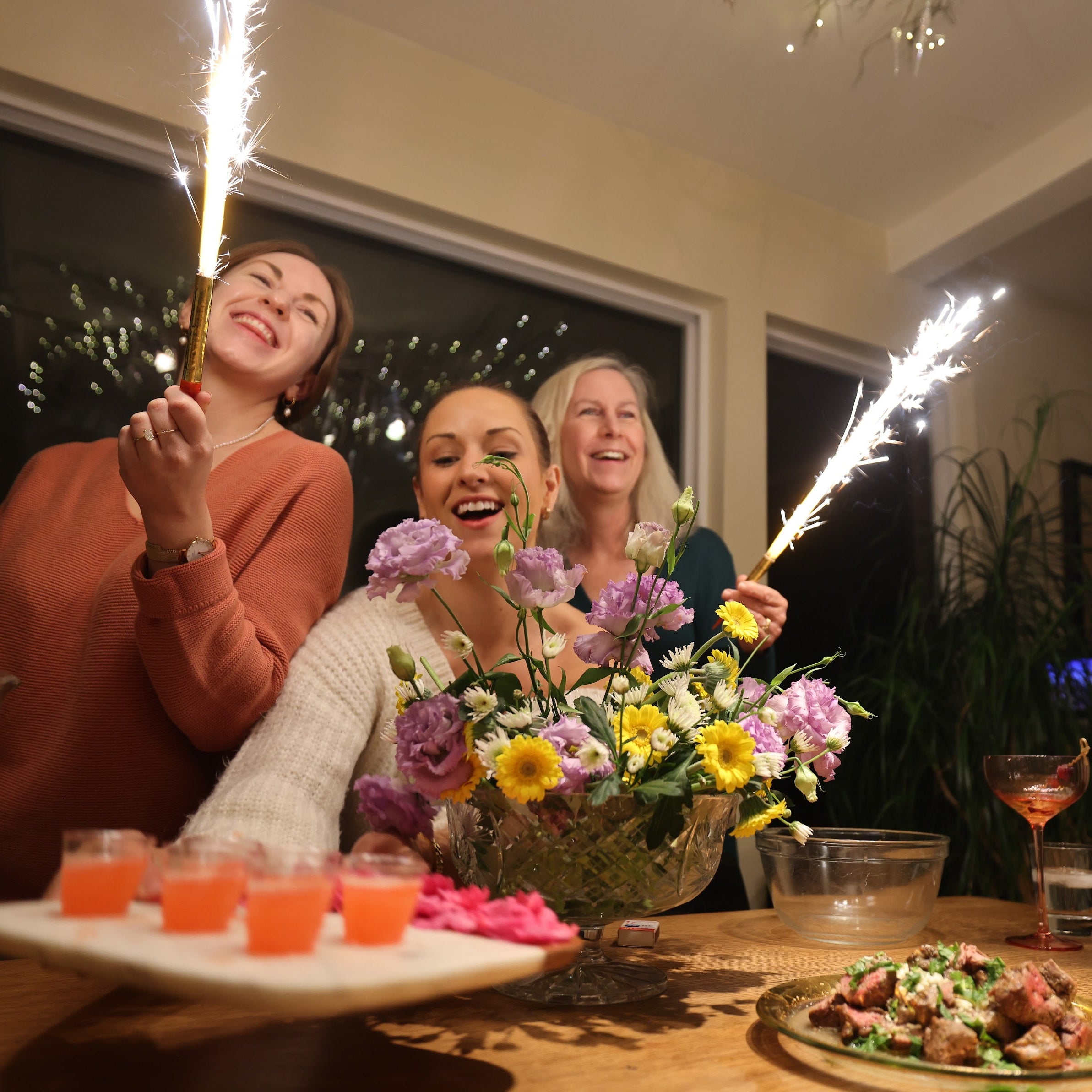 Three women celebrating with sparklers and a floral arrangement at a dinner table.