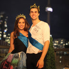 Two people wearing crowns and sashes against a city skyline at night.