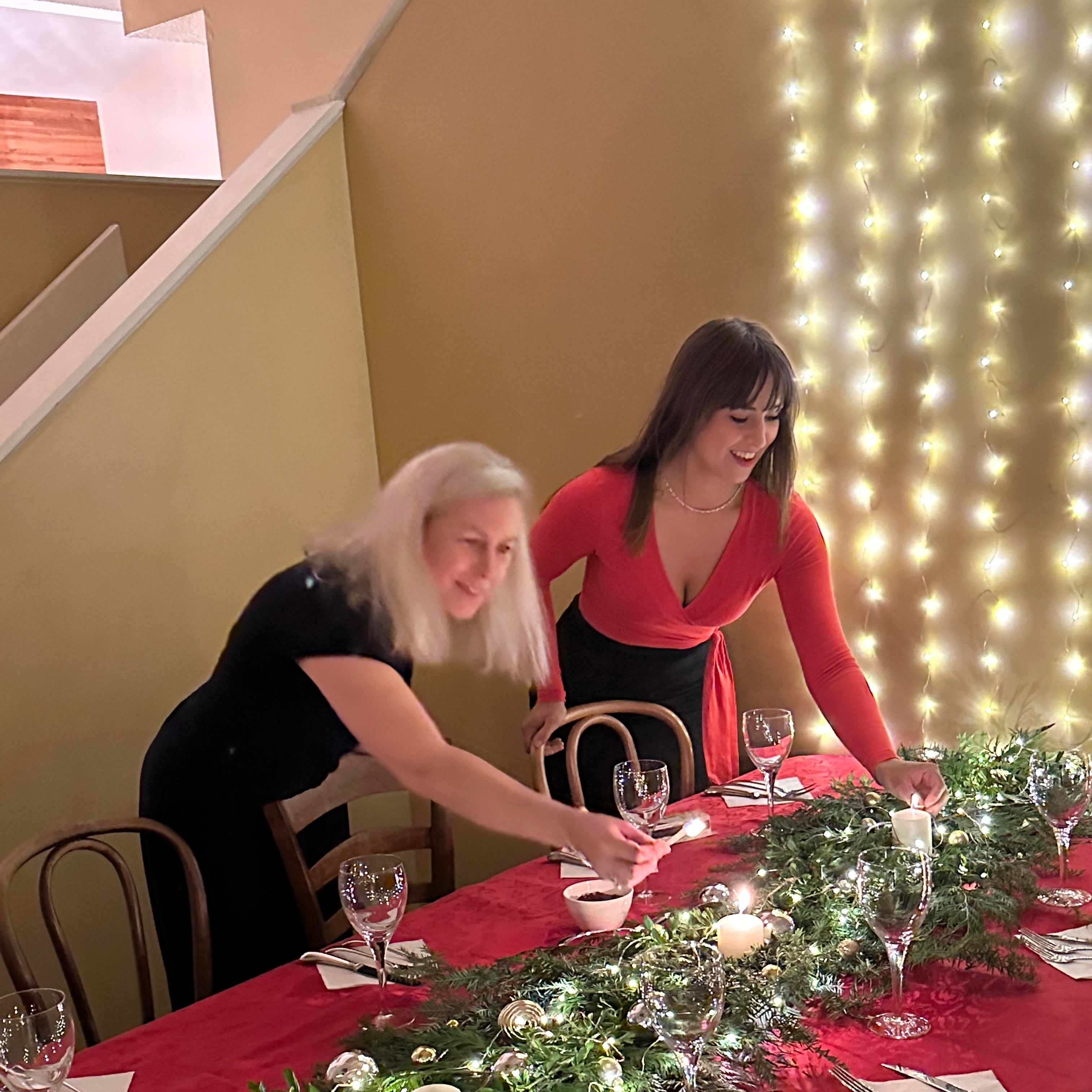 Two women setting a festive table with Christmas decorations and lights.