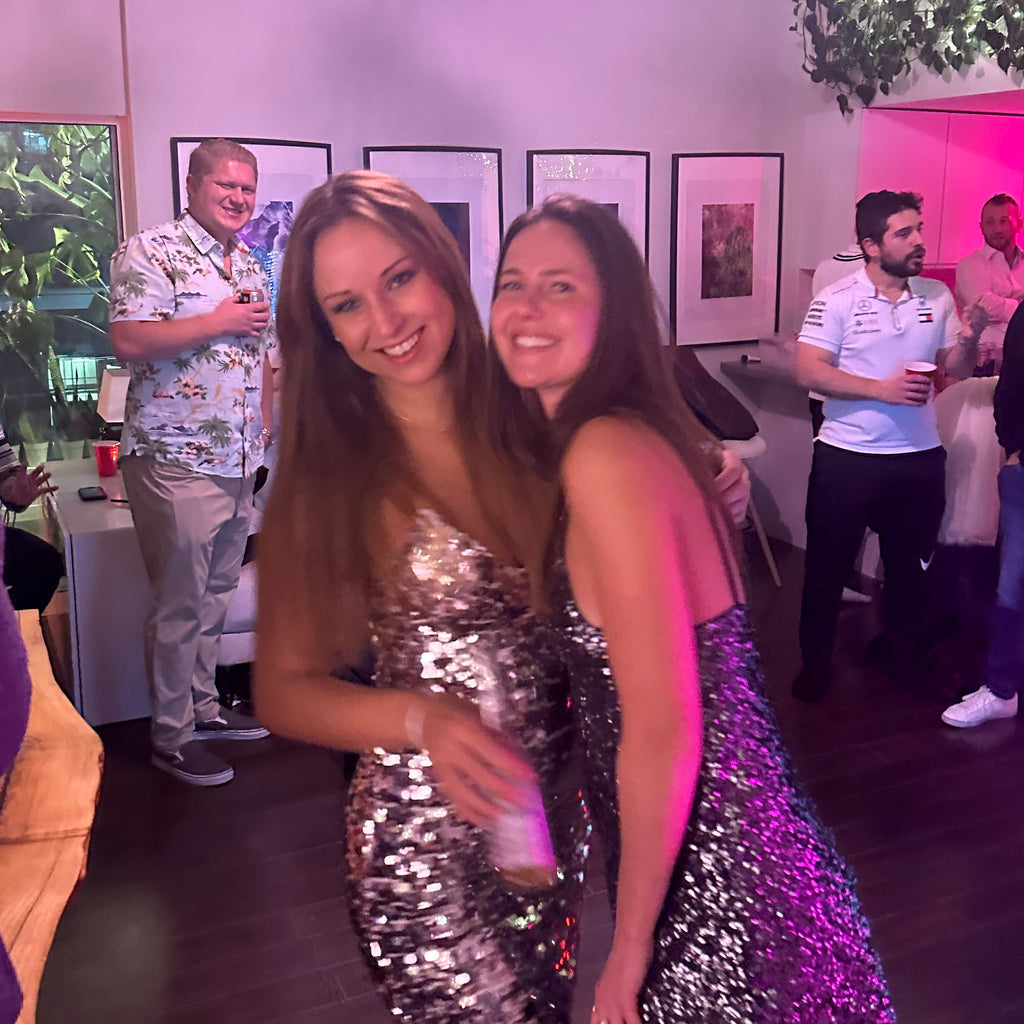 Two women in sparkly dresses posing together at a social event with people in the background.