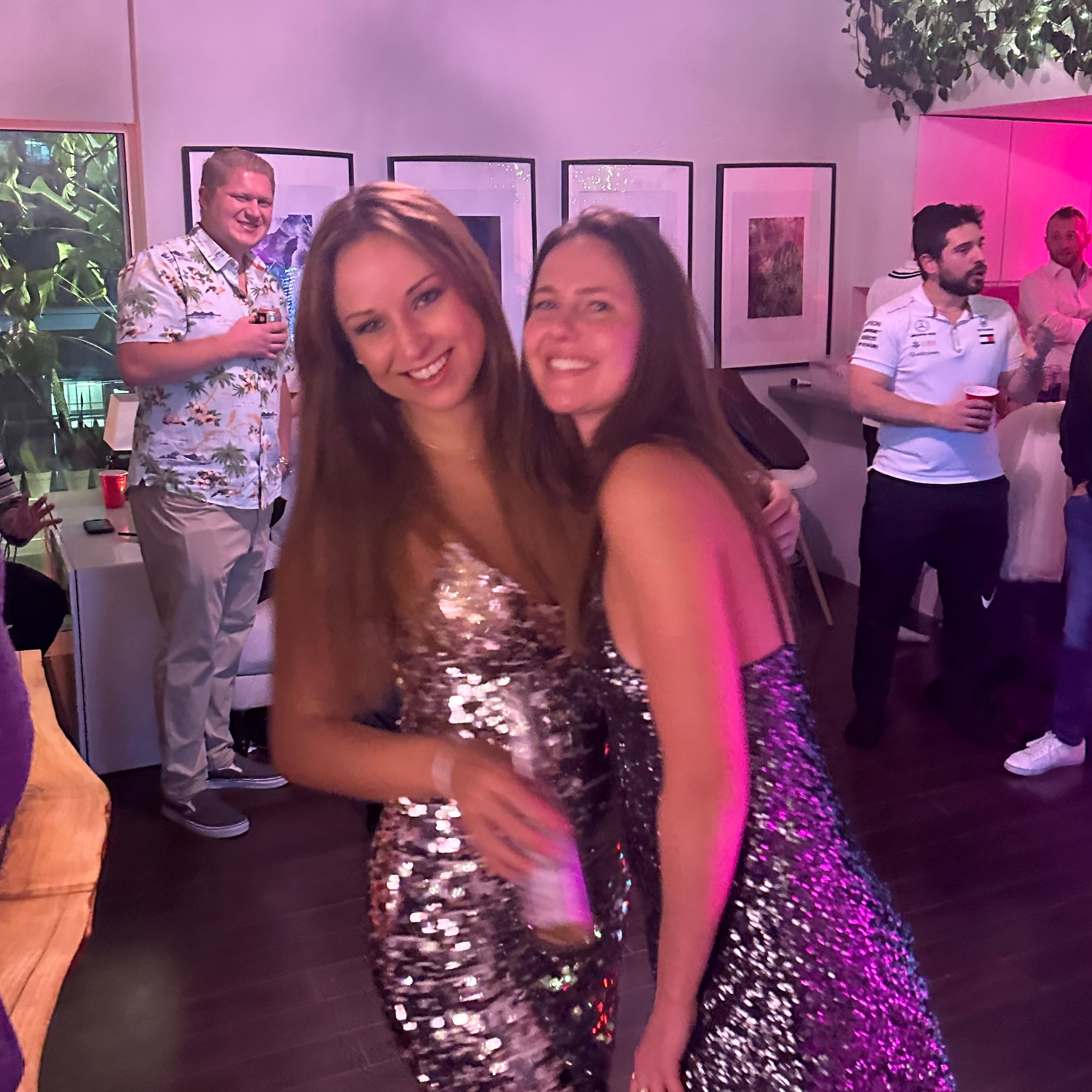 Two women in sparkly dresses posing together at a social event with people in the background.