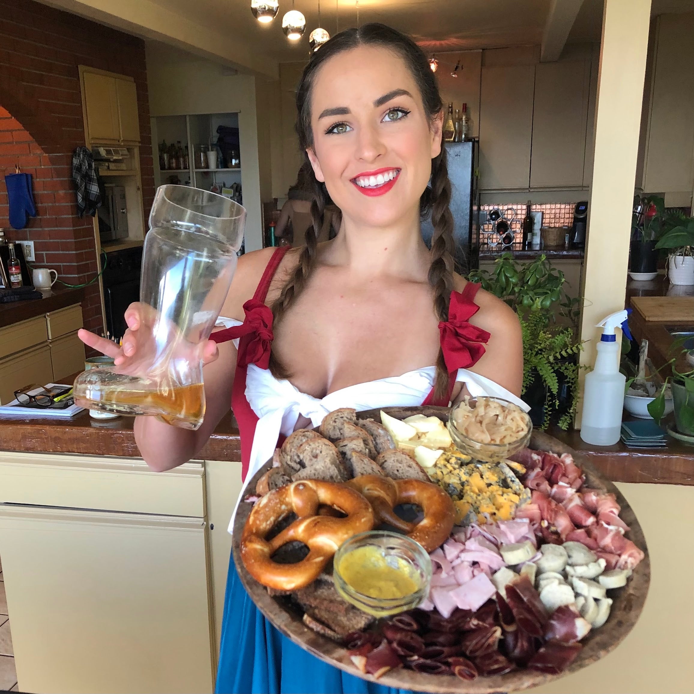 Woman holding a large platter of food and a glass of beer in a kitchen.