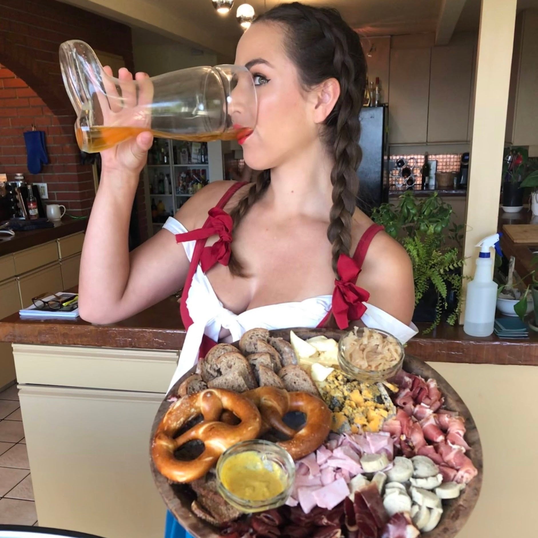 Woman holding a large platter of food and a glass of beer in a kitchen setting.