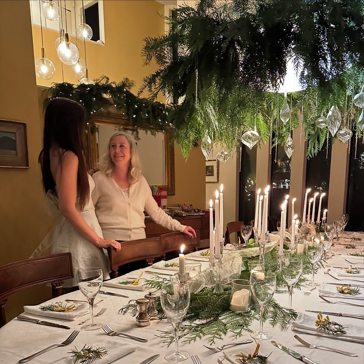 Woman setting a festive dining table with candles and greenery, in a home setting.
