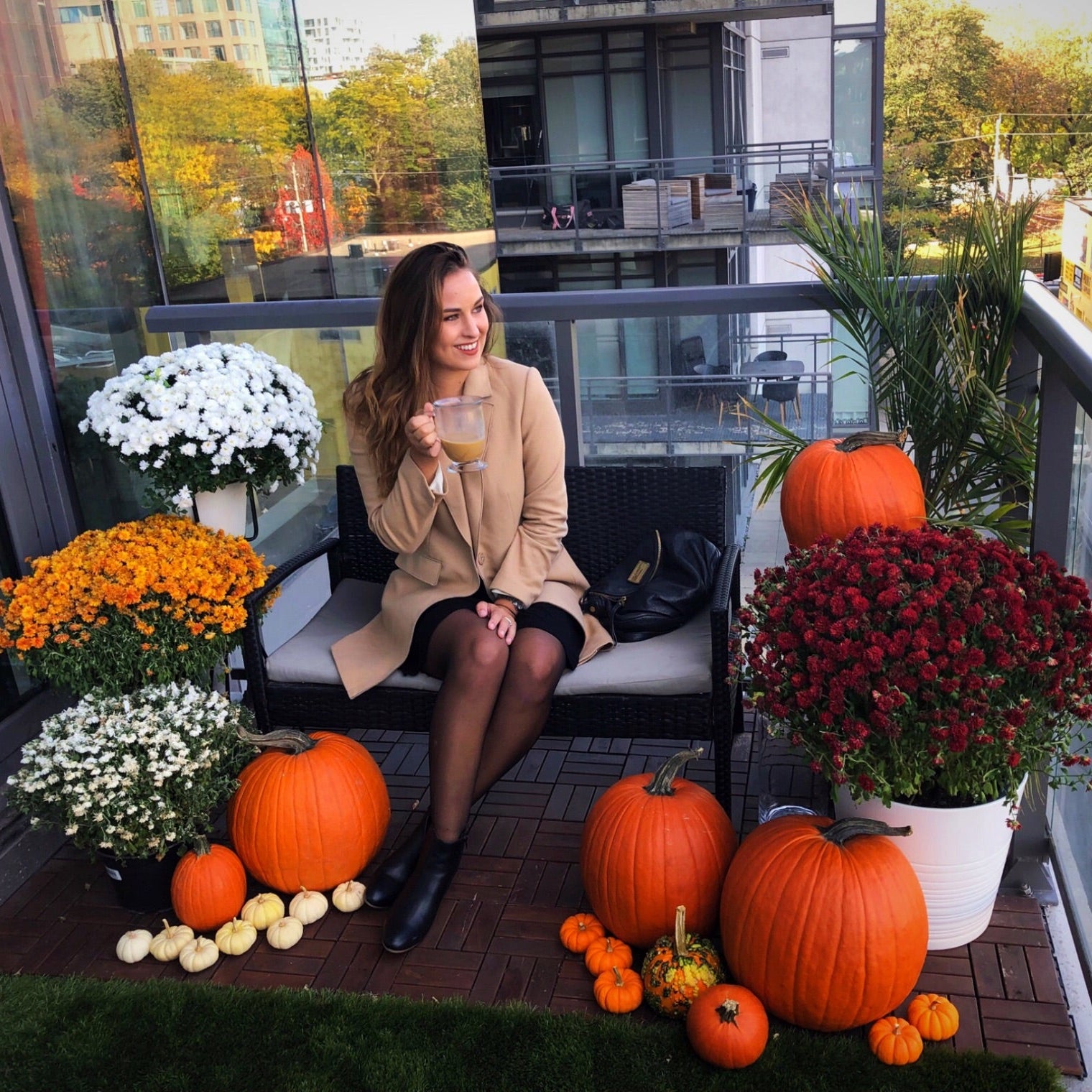 Woman sitting on a balcony with pumpkins and flowers, surrounded by urban scenery.