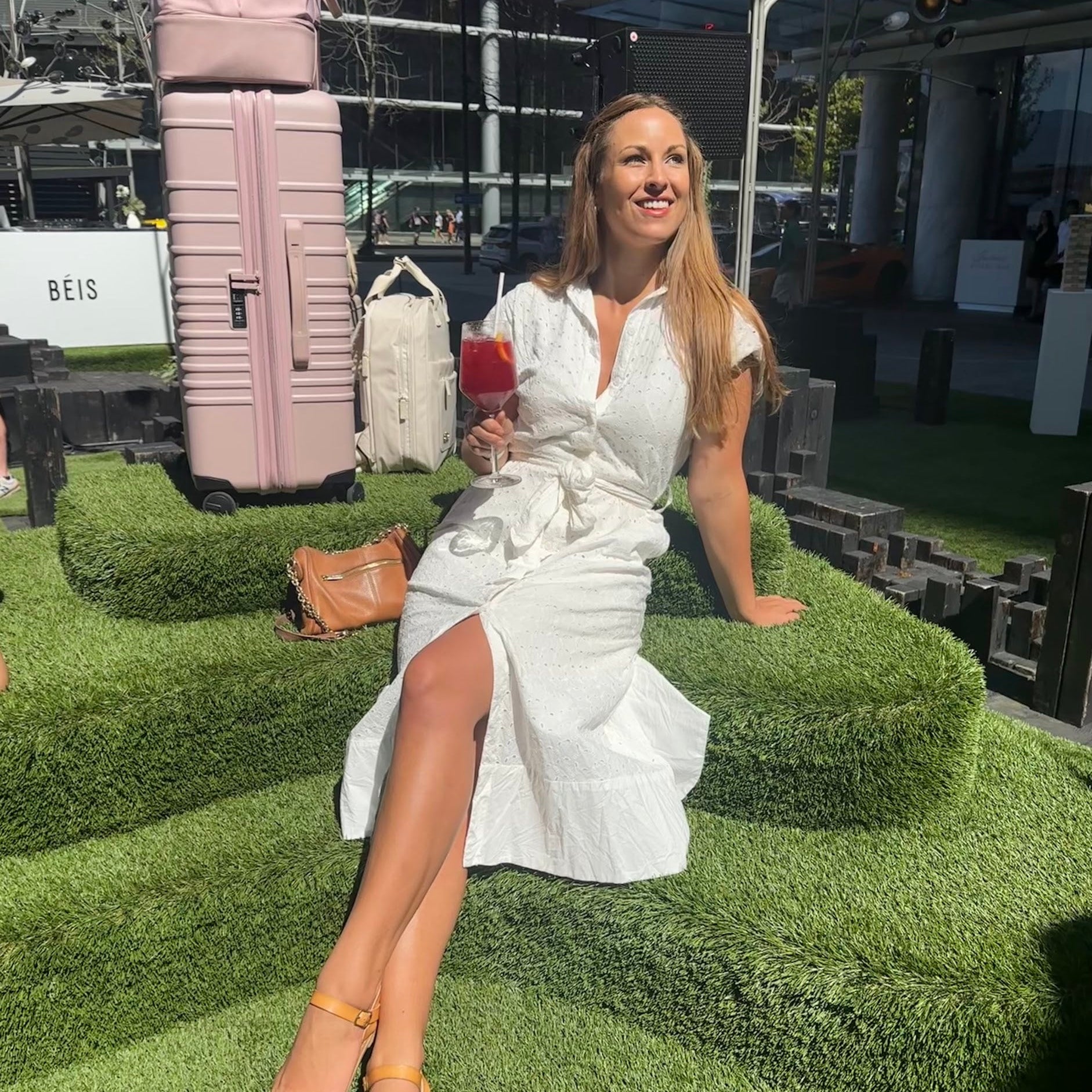 Woman in a white dress sitting on grass with bags and a drink in the background
