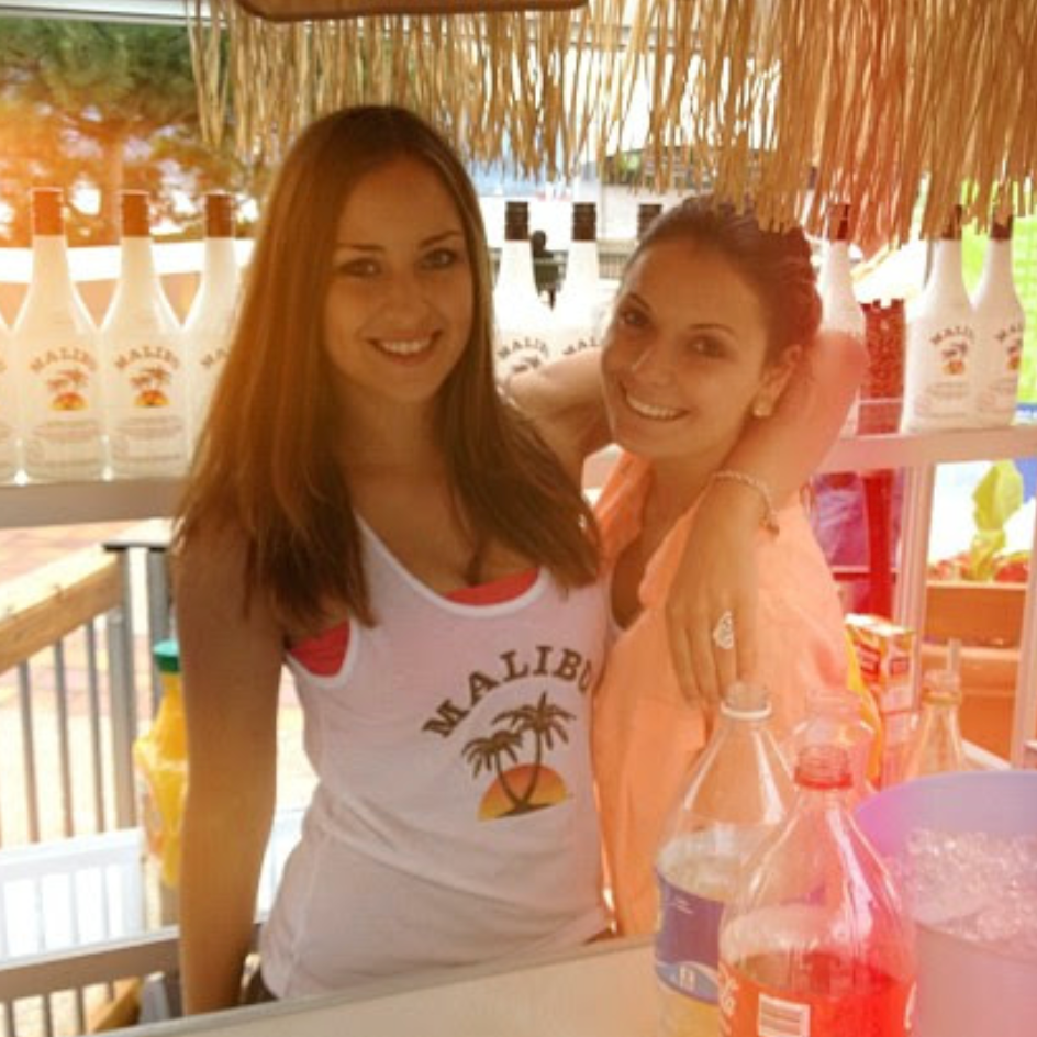 Two women standing behind a bar with drinks and a Malibu bottle in the background.
